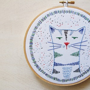 May include: Embroidered hoop art featuring a stylized blue and green cat with a red nose. The cat is surrounded by a colorful border of blue, green, red, and black stitches.