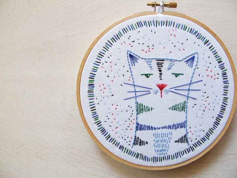 May include: Embroidered hoop art featuring a stylized blue and green cat with a red nose. The cat is surrounded by a colorful border of blue, green, red, and black stitches.