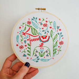 May include: Embroidered hoop art featuring a stylized red and blue Dala horse with green foliage and red and blue flowers. The design is surrounded by a white background and a wooden hoop.