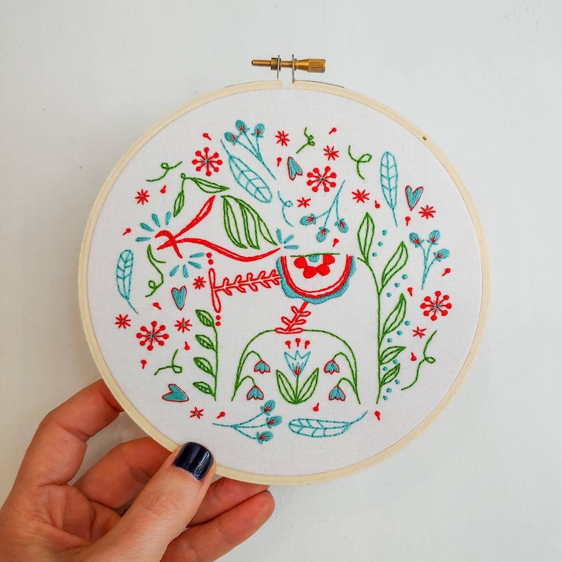 May include: Embroidered hoop art featuring a stylized red and blue Dala horse with green foliage and red and blue flowers. The design is surrounded by a white background and a wooden hoop.