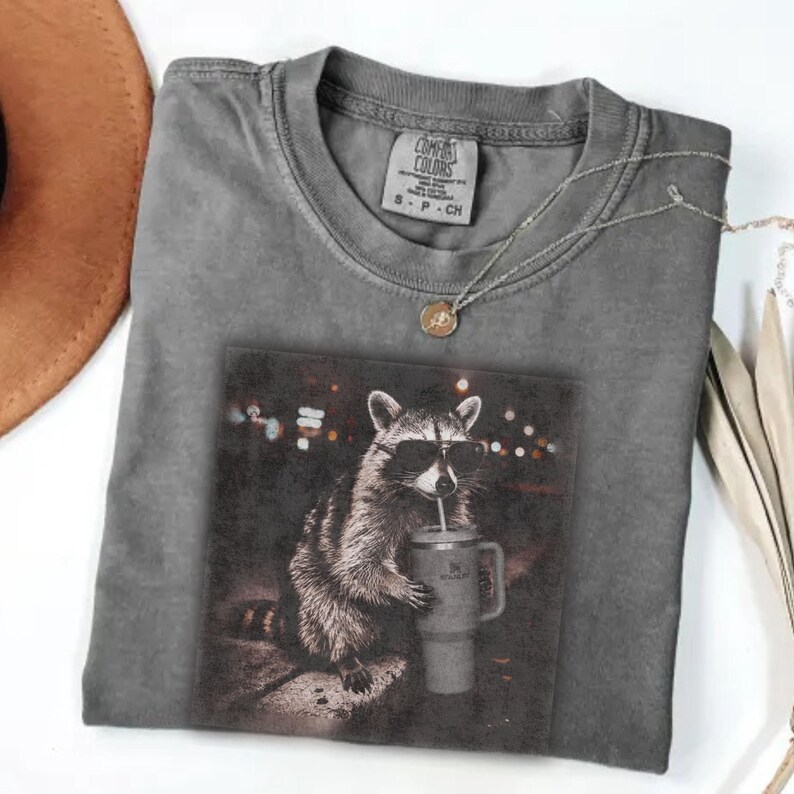 May include: A grey Comfort Colors t-shirt with a graphic of a raccoon wearing sunglasses and drinking from a tumbler. The shirt has a crew neck and short sleeves. A silver necklace with a pendant is also visible.