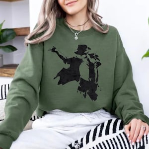 May include: An olive green long-sleeve sweatshirt featuring a black graphic print of a person in a fedora hat and suit performing a dance pose. The garment has a crew neckline and long sleeves, with a distressed ink-blot style illustration.