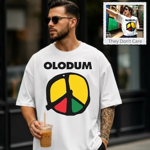May include: A white short-sleeved t-shirt featuring the black text OLODUM above a large circular peace sign graphic. The peace sign is divided into three sections colored yellow, red, and green with thick black outlines. The shirt has a crew neckline.
