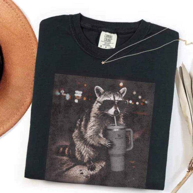 May include: Black Comfort Colors t-shirt with a raccoon wearing sunglasses and holding a Stanley tumbler. The graphic shows a raccoon with a drink against a blurred city lights background. A gold necklace with a star pendant is on the shirt.