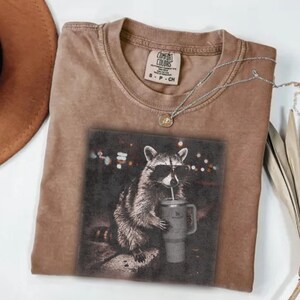 May include: A brown Comfort Colors t-shirt with a graphic of a raccoon wearing sunglasses and drinking from a tumbler. The shirt has a crew neck and short sleeves. A silver necklace with a small pendant is also visible.