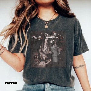 May include: Dark grey t-shirt with a raccoon wearing sunglasses and holding a drink. The raccoon graphic is in a square frame with a blurred city lights background. The word "PEPPER" is printed at the bottom of the shirt.