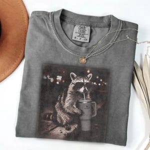 May include: Grey Comfort Colors t-shirt with a graphic of a raccoon wearing sunglasses and drinking from a tumbler. The shirt has a crew neck and a small tag near the collar. A silver necklace and a brown hat are also visible.