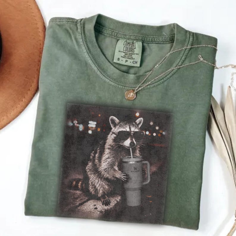 May include: Green Comfort Colors t-shirt with a graphic of a raccoon wearing sunglasses and drinking from a tumbler. A gold necklace and a brown hat are also visible. The shirt has a small tag at the neckline.