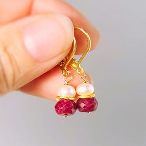 May include: Gold-colored dangle earrings with a pearl and ruby design. Each earring features a small, round, white pearl above a faceted, deep red ruby. The earrings are held up by gold-colored hooks.