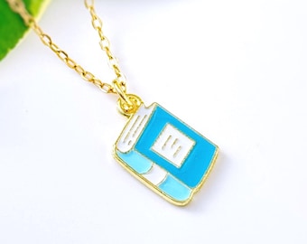 May include: A gold-colored necklace with a pendant shaped like a small book. The book pendant is blue and white with gold trim. The necklace is displayed against a white background with a green leaf.