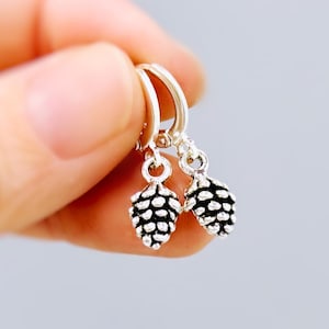 May include: Silver hoop earrings with small, detailed pinecone charms. The earrings are held against a soft, blurred background. The pinecone charms have a textured surface, adding visual interest to the jewelry.