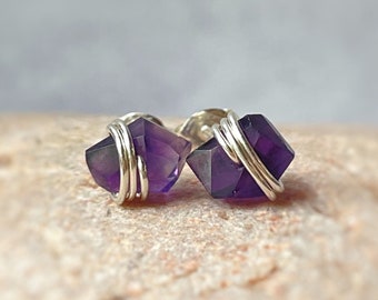 May include: A pair of stud earrings featuring deep purple, faceted amethyst crystals. Each crystal is secured with a silver-colored wire wrap that encircles the center of the stone, creating a distinct, geometric appearance for these small accessories.