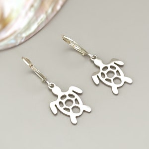 May include: A pair of silver-toned dangle earrings featuring a cutout sea turtle design. Each earring is suspended from a leverback closure, showcasing a detailed shell pattern with circular and oval cutouts within the turtle's body shape.