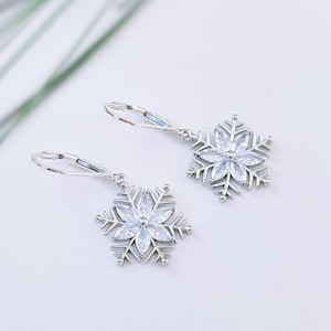 May include: A pair of silver snowflake earrings. Each earring features a snowflake design with clear, sparkling crystals. The earrings have a lever-back closure. The background is white with a hint of green.
