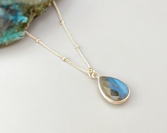 May include: A silver chain necklace with a teardrop-shaped labradorite gemstone pendant. The gemstone is a deep blue with flashes of green and gold.