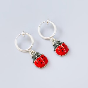 May include: A pair of silver hoop earrings featuring dangling ladybug charms. Each charm is painted with a bright red body, black spots, and a black head. The earrings have a simple circular hoop design with a small metal loop connecting the ladybug.