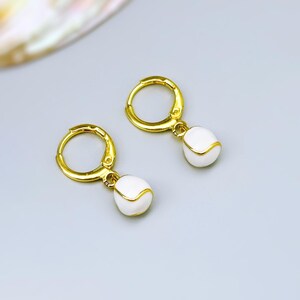 May include: A pair of gold hoop earrings featuring small, white, rounded charms. Each charm has a smooth, glossy finish with a thin gold line accenting the surface. The earrings are designed with a hinged clasp mechanism for secure closure.