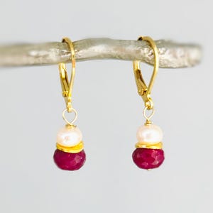 May include: A pair of gold-colored leverback earrings. Each earring features a white pearl, a gold-colored spacer, and a faceted, deep red gemstone. The earrings are suspended from a silver-colored branch.