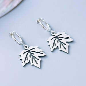 May include: A pair of silver-toned hoop earrings, each featuring a dangling charm shaped like a maple leaf. The leaf charms have a cut-out design that highlights the veins of the leaf, and the hoops are circular with a simple, polished finish.