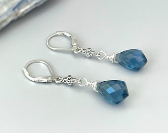 May include: A pair of silver dangle earrings with blue gemstone drops. The earrings have a delicate silver chain with a small flower design.