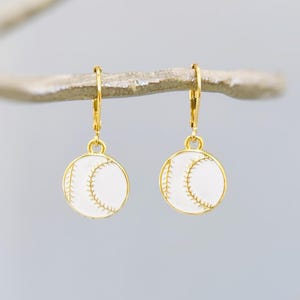 May include: A pair of gold-toned leverback earrings featuring circular charms designed to resemble white baseballs with gold-coloured stitching details. The earrings have a polished finish and a simple, round shape that hangs from the ear wire.
