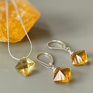 May include: A set of gold and silver jewelry featuring a necklace and earrings. The necklace has a delicate chain and a square-shaped pendant with a faceted gold gemstone. The earrings have silver hooks and dangle square-shaped pendants with faceted gold gemstones.