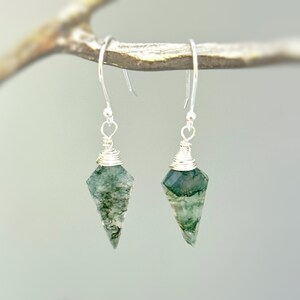 May include: A pair of dangle earrings featuring faceted, triangular moss agate stones with green inclusions. Each stone is secured with silver-colored wire wrapping at the top and attached to a simple silver-colored metal hook for ear suspension.