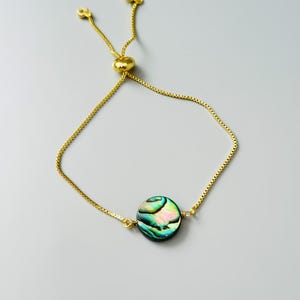 May include: A gold-toned adjustable bracelet featuring a circular abalone shell charm. The bracelet has a delicate box chain design with a sliding bead mechanism for sizing, and the central shell displays iridescent green, blue, and pink hues.
