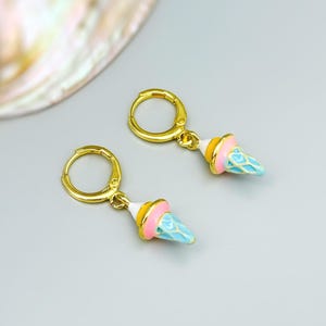 May include: A pair of gold hoop earrings featuring dangling ice cream cone charms. Each charm is detailed with enamel in shades of light blue, pink, and yellow, depicting a textured cone and a rounded scoop of ice cream on a gold metal base.