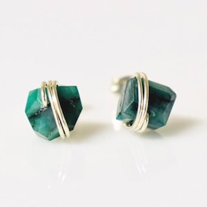 May include: A pair of stud earrings featuring raw, deep green malachite stones. Each stone is secured by a silver-toned metal wire wrap that encircles the centre of the gem. The earrings have a polished, metallic post backing for secure attachment.