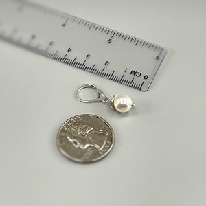 Puede incluir: Un pendiente de plata con cierre de palanca y una perla blanca redonda. El pendiente est&aacute; colocado junto a una moneda de cuarto de d&oacute;lar estadounidense y una regla de pl&aacute;stico transparente que muestra una longitud de unos 2 cm.