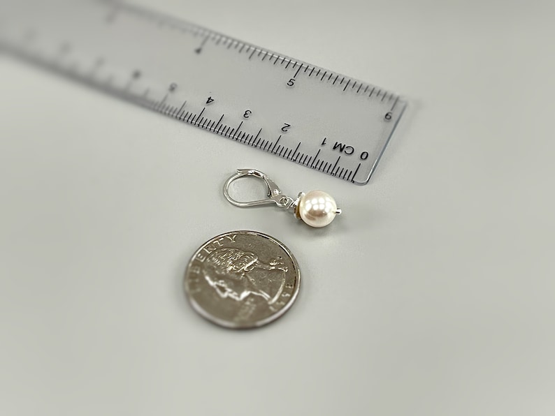 Puede incluir: Un pendiente de plata con cierre de palanca y una perla blanca redonda. El pendiente est&aacute; colocado junto a una moneda de cuarto de d&oacute;lar estadounidense y una regla de pl&aacute;stico transparente que muestra una longitud de unos 2 cm.