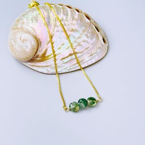 May include: A gold-toned chain necklace featuring three small, round, faceted green moss agate beads. The necklace is draped over a large, iridescent abalone shell that displays swirling shades of pink, cream, and pearlescent white tones.