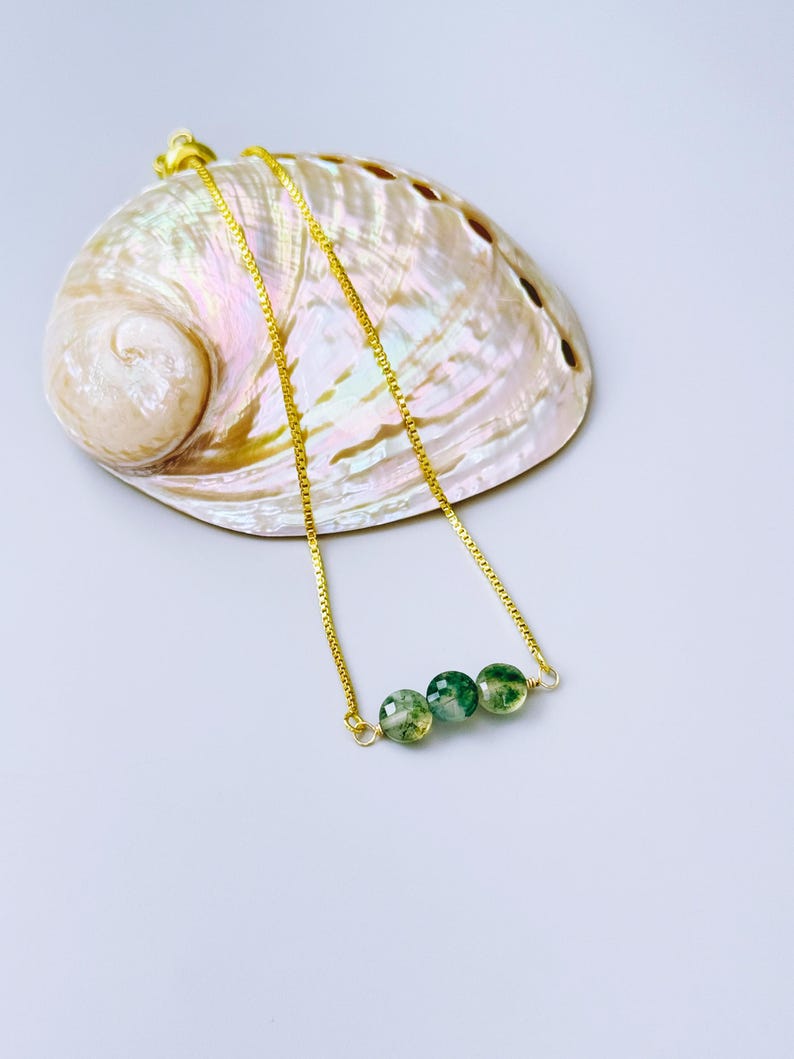 May include: A gold-toned chain necklace featuring three small, round, faceted green moss agate beads. The necklace is draped over a large, iridescent abalone shell that displays swirling shades of pink, cream, and pearlescent white tones.