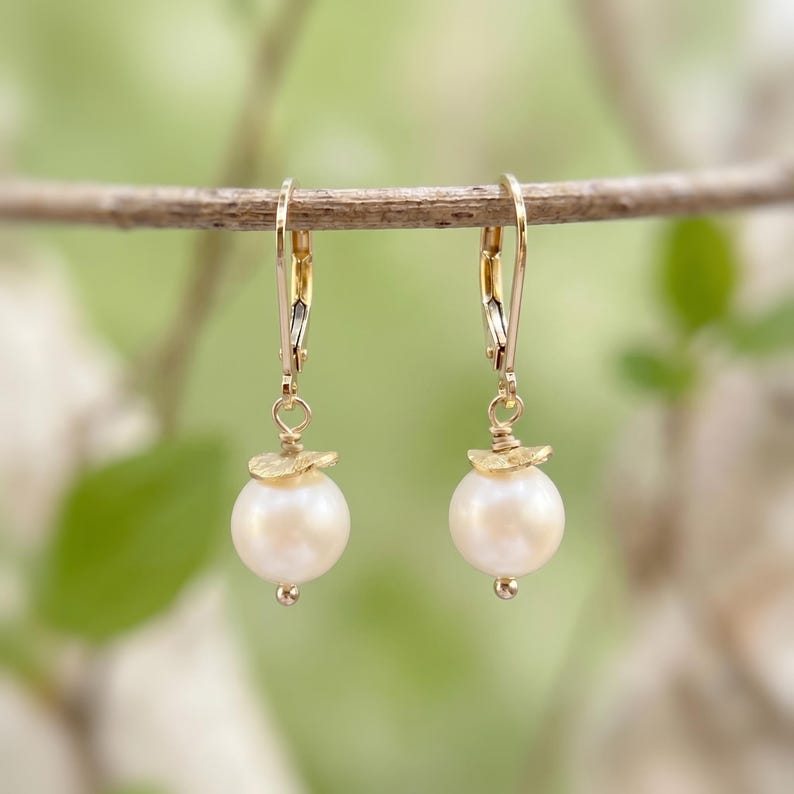 Puede incluir: Un par de pendientes colgantes con perlas blancas redondas suspendidas de ganchos dorados tipo dormilona. Cada perla est&aacute; rematada con un peque&ntilde;o disco de metal dorado texturizado y una diminuta cuenta en la base del dise&ntilde;o.