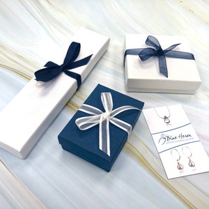 May include: Three gift boxes with blue and white ribbon. The boxes are on a marble surface. The text on the card reads "Blue Heron Jewelry & Hair Accessories."