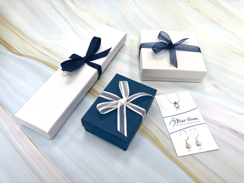 May include: Three gift boxes with blue and white ribbon. The boxes are on a marble surface. The text on the card reads "Blue Heron Jewelry & Hair Accessories."