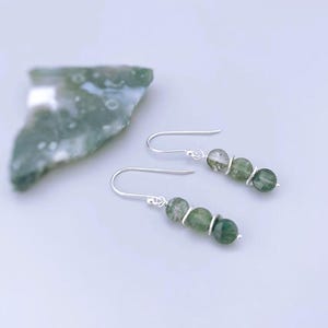May include: A pair of drop earrings featuring three round, variegated green moss agate beads separated by thin silver spacers, suspended from simple silver ear wires. The beads display natural inclusions and varying shades of forest green.