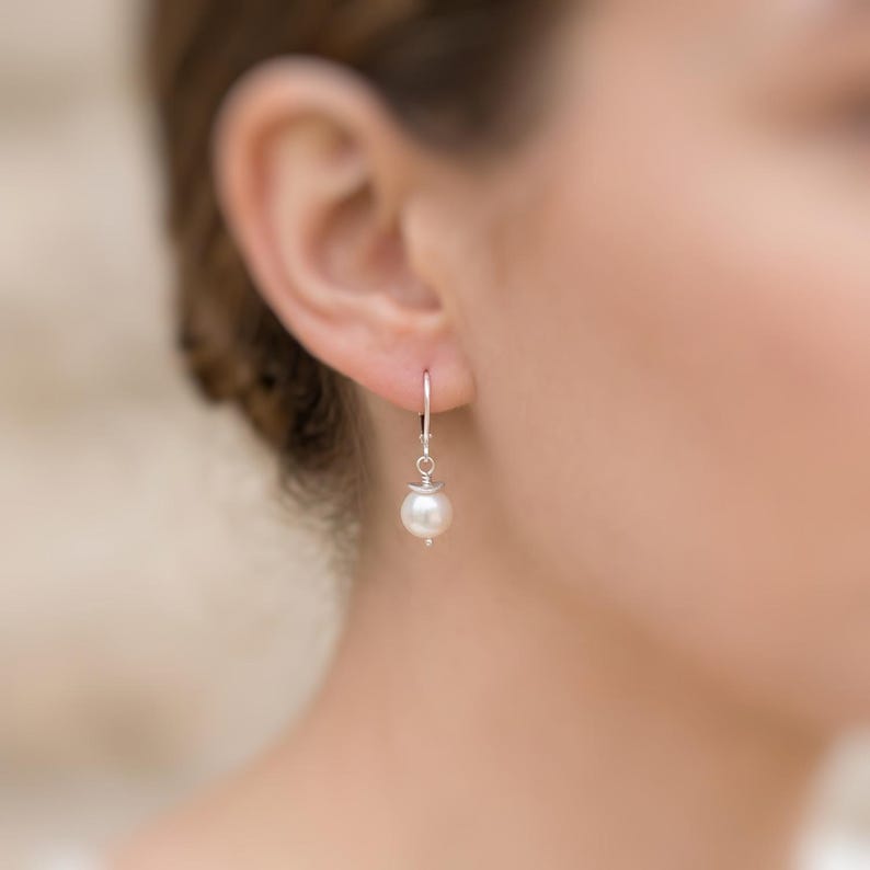 Puede incluir: Un pendiente colgante individual que presenta una perla blanca redonda suspendida de un fino gancho de plata. La perla est&aacute; unida al alambre de metal mediante una peque&ntilde;a tapa de plata, creando un dise&ntilde;o simple y limpio que cuelga verticalmente.