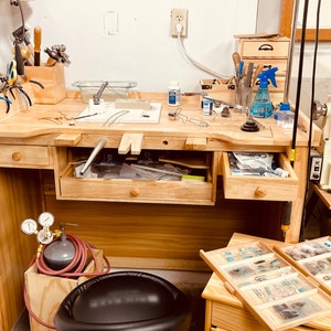 Puede incluir: Un banco de trabajo de joyero de madera clara con una abertura central, varios cajones y una superficie superior organizada con diversas herramientas de metal, alicates, una estaci&oacute;n de soldadura, un pulverizador azul y una peque&ntilde;a caja organizadora.