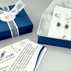 May include: A pair of silver-toned leverback earrings featuring dark, spherical beads, presented on a white display card inside a sheer organza bag, alongside a blue gift box with a white ribbon and a care instruction card with a heron logo.