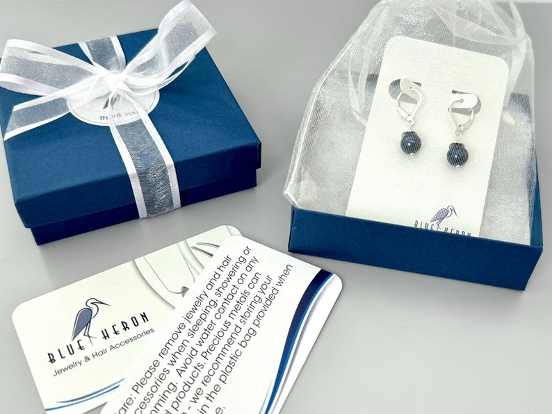 May include: A pair of silver-toned leverback earrings featuring dark, spherical beads, presented on a white display card inside a sheer organza bag, alongside a blue gift box with a white ribbon and a care instruction card with a heron logo.
