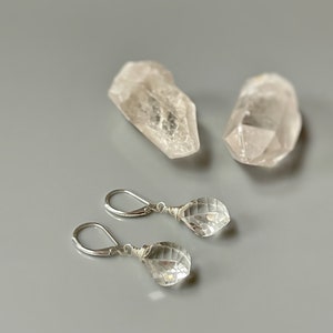 May include: A pair of silver earrings with clear quartz crystal drops. The earrings are on a gray surface with two clear quartz crystals.