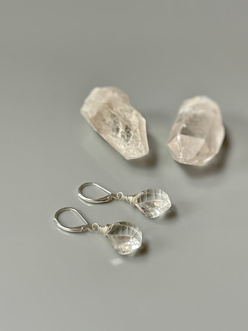 May include: A pair of silver earrings with clear quartz crystal drops. The earrings are on a gray surface with two clear quartz crystals.