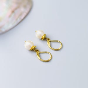 Puede incluir: Un par de pendientes con cierre de palanca en tono dorado, cada uno con una perla blanca redonda suspendida de una peque&ntilde;a tapa de metal dorado. Los pendientes tienen un acabado pulido y un dise&ntilde;o colgante cl&aacute;sico y sencillo.