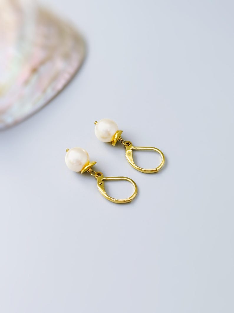 Puede incluir: Un par de pendientes con cierre de palanca en tono dorado, cada uno con una perla blanca redonda suspendida de una peque&ntilde;a tapa de metal dorado. Los pendientes tienen un acabado pulido y un dise&ntilde;o colgante cl&aacute;sico y sencillo.
