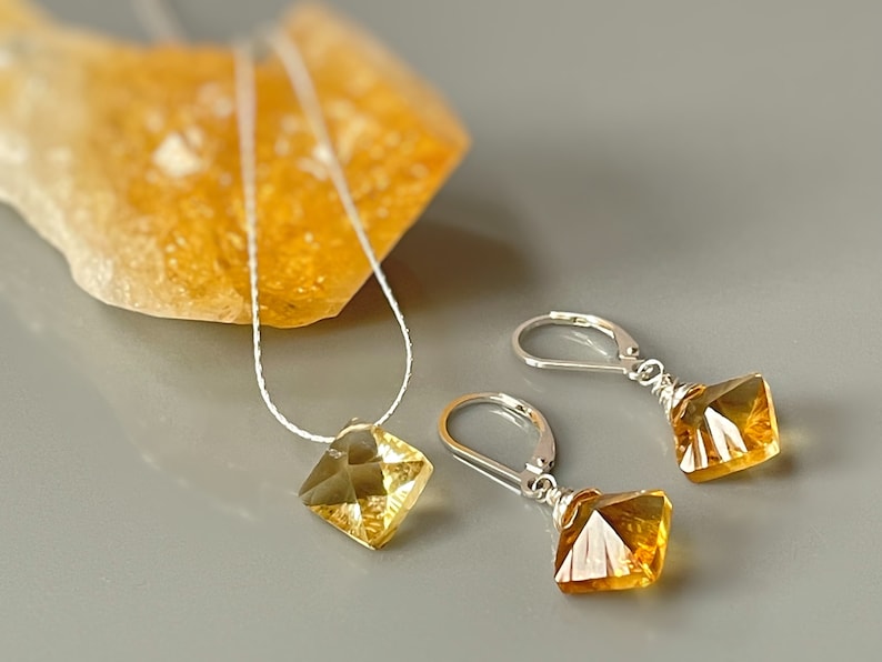 May include: A set of gold and silver jewelry featuring a necklace and earrings. The necklace has a delicate chain and a square-shaped pendant with a faceted gold gemstone. The earrings have silver hooks and dangle square-shaped pendants with faceted gold gemstones.