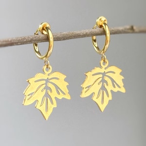 May include: A pair of gold-toned hoop earrings featuring dangling, cut-out leaf charms. Each earring consists of a smooth, circular hoop connected to a flat, stylized maple leaf pendant with intricate, open-work vein detailing throughout.