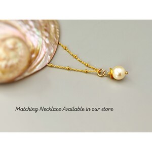 Puede incluir: Un collar de cadena dorada con peque&ntilde;as cuentas doradas espaciadas uniformemente y un colgante de perla de color crema. El texto "Matching Necklace Available in our store" est&aacute; impreso debajo de la joya. El collar descansa cerca de una concha.
