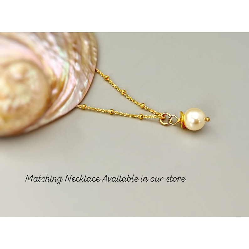 Puede incluir: Un collar de cadena dorada con peque&ntilde;as cuentas doradas espaciadas uniformemente y un colgante de perla de color crema. El texto "Matching Necklace Available in our store" est&aacute; impreso debajo de la joya. El collar descansa cerca de una concha.